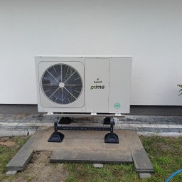 WODMAT - Zewnętrzna jednostka klimatyzacji marki Galmet Prima, zamontowana na betonowej podstawie w Zamościu, obok mała choinka w czarnej doniczce, na tle białej ściany budynku.