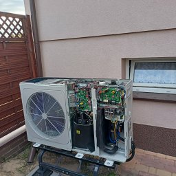 WODMAT - Otwarta obudowa jednostki zewnętrznej urządzenia grzewczego na metalowym stojaku, z widocznymi elementami elektroniki i wentylatorem, na tle ściany budynku z oknem i drewnianego płotu.