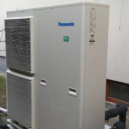 WODMAT - Zewnętrzna jednostka pompy ciepła Panasonic R32 zamontowana na betonowej podstawie z drewnianym obramowaniem, z widocznymi rurami w izolacji termicznej i oknem w tle.