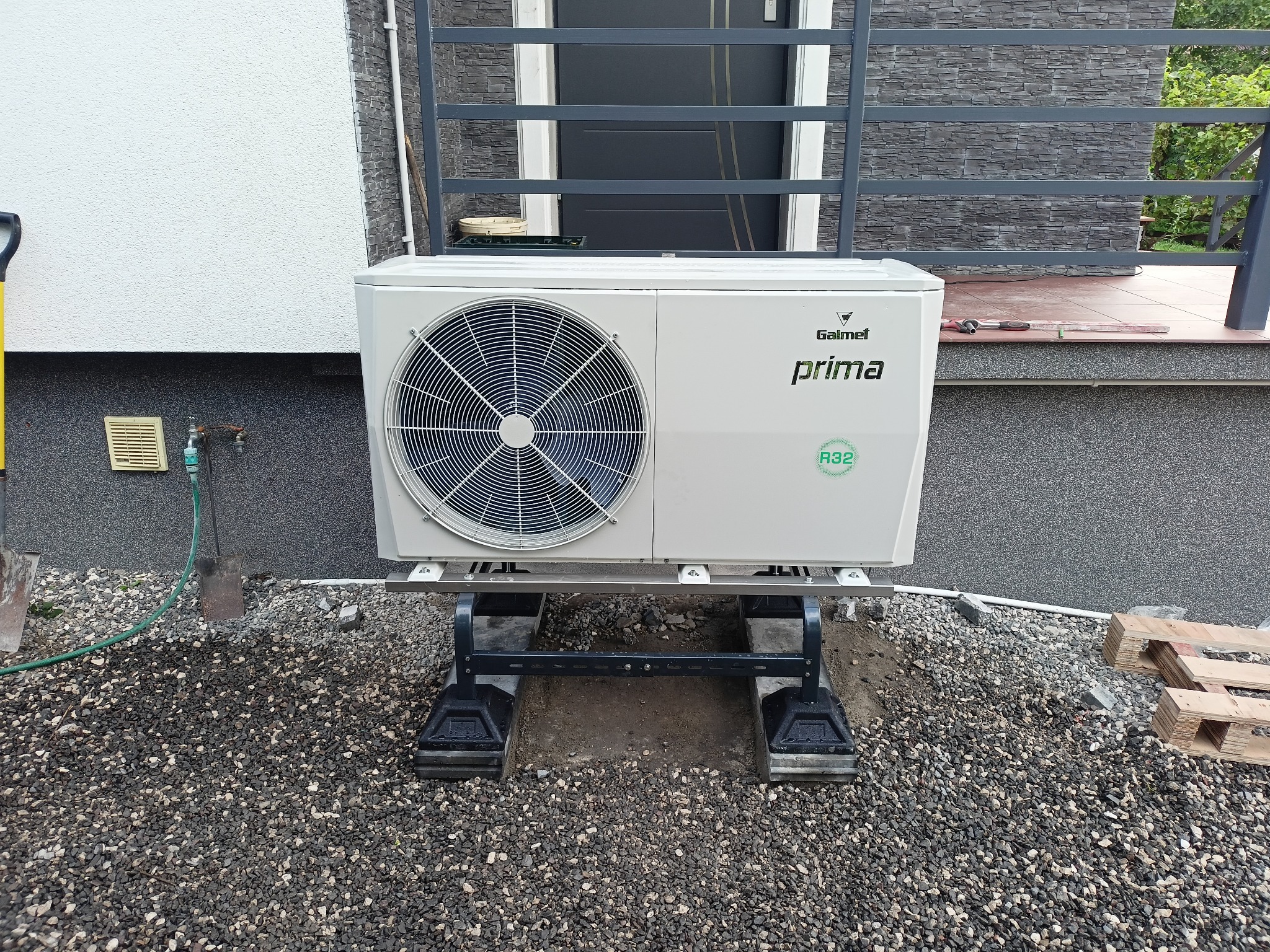Biała jednostka zewnętrzna pompy ciepła Galmet Prima R32 zamontowana na stalowej konstrukcji na żwirowym podłożu, widoczna łopata i wąż ogrodowy, montaż w toku.