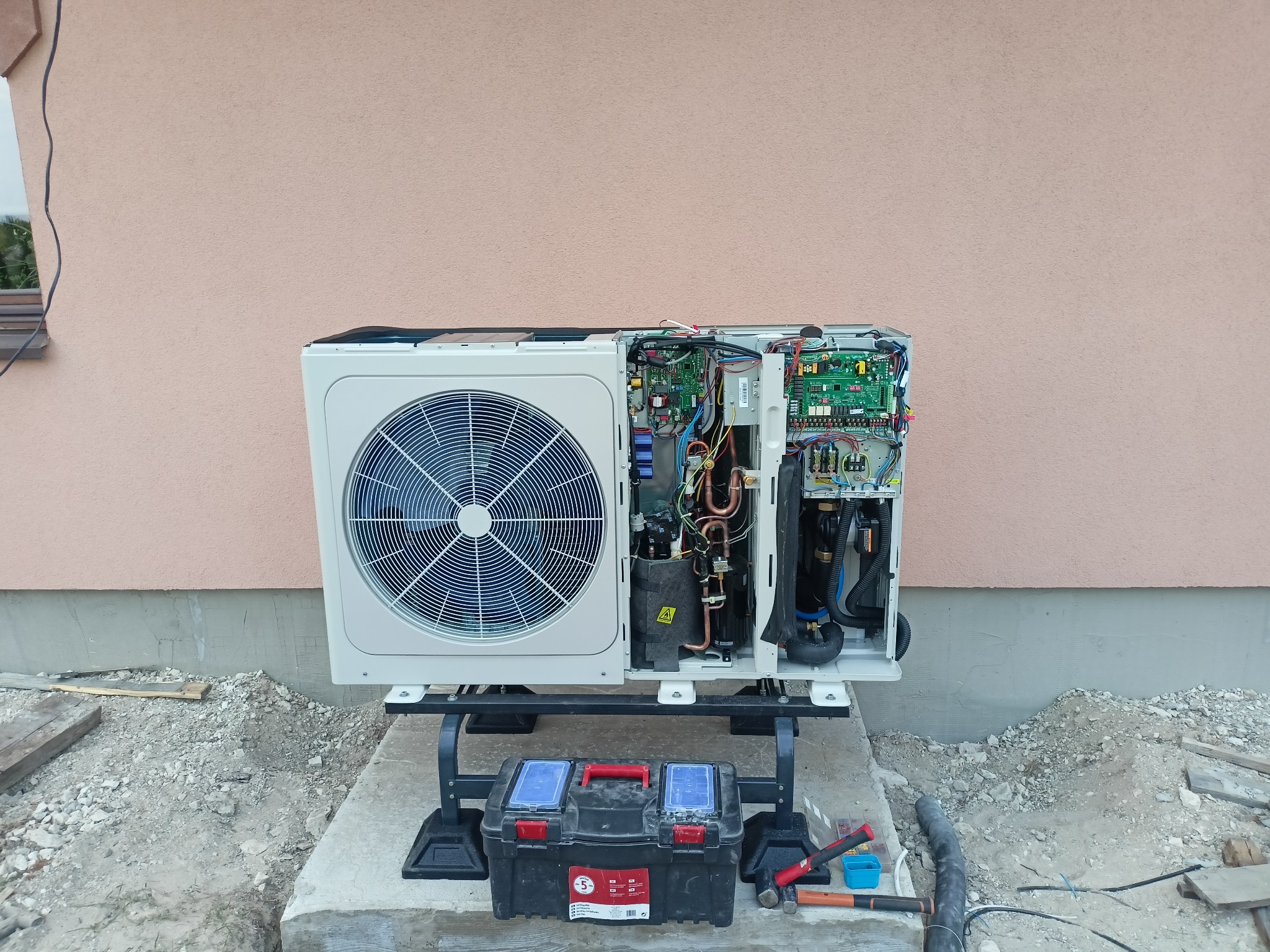Otwarta obudowa zewnętrznej jednostki pompy ciepła podczas montażu, widoczne wewnętrzne komponenty elektryczne i hydrauliczne, skrzynka z narzędziami na betonowym podłożu, różowa ściana budynku w tle.