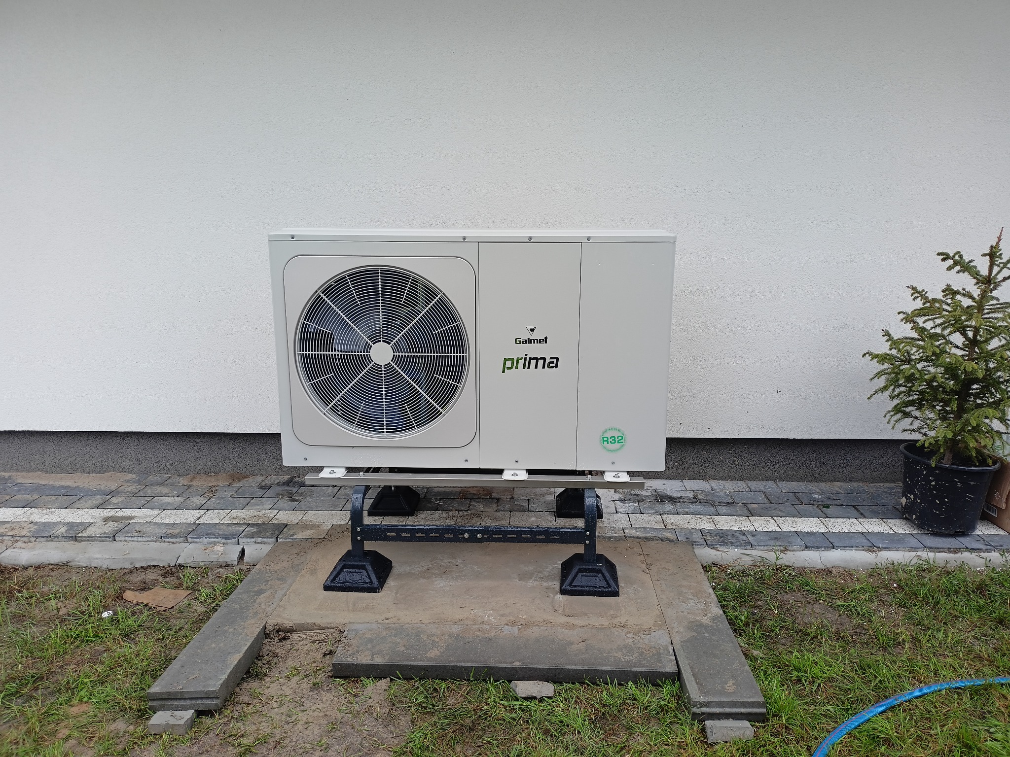 Zewnętrzna jednostka klimatyzacji marki Galmet Prima, zamontowana na betonowej podstawie w Zamościu, obok mała choinka w czarnej doniczce, na tle białej ściany budynku.