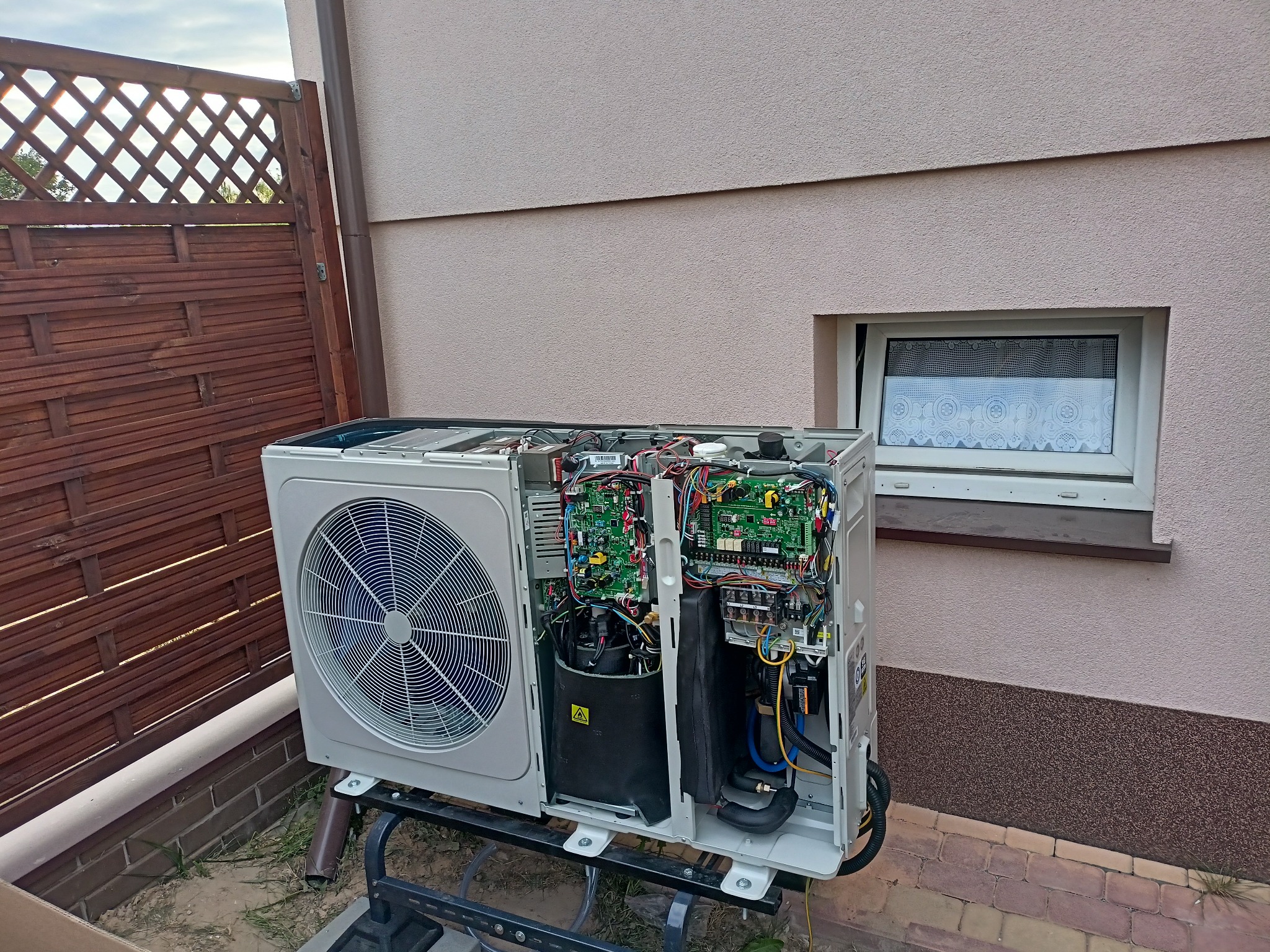 Otwarta obudowa jednostki zewnętrznej urządzenia grzewczego na metalowym stojaku, z widocznymi elementami elektroniki i wentylatorem, na tle ściany budynku z oknem i drewnianego płotu.