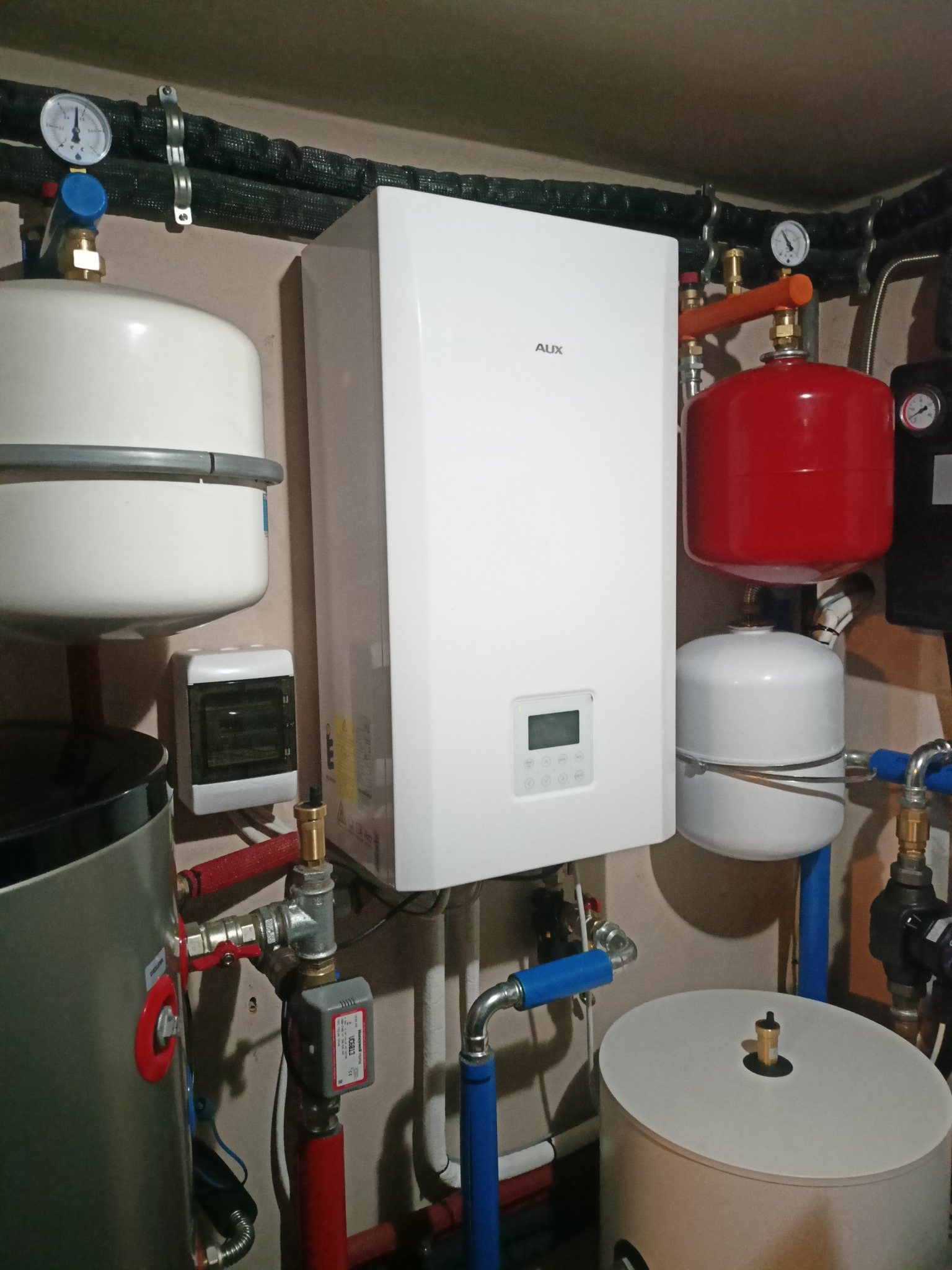 Biały piec gazowy marki AUX zamontowany na ścianie, z widocznymi elementami instalacji grzewczej: zbiorniki wyrównawcze białe i czerwony, rury miedziane i plastikowe, manometry i zawory.