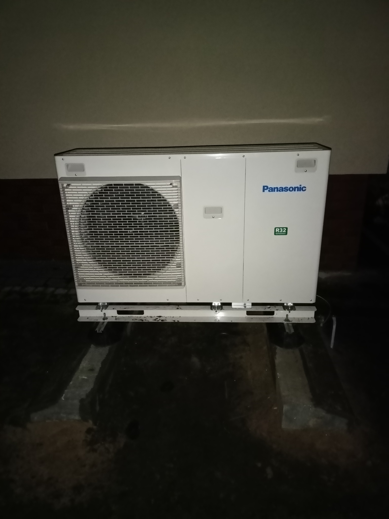 Biała zewnętrzna jednostka marki Panasonic z widoczną kratką wentylatora i oznaczeniem czynnika chłodniczego R32, zamontowana na metalowej konstrukcji w słabo oświetlonym pomieszczeniu.