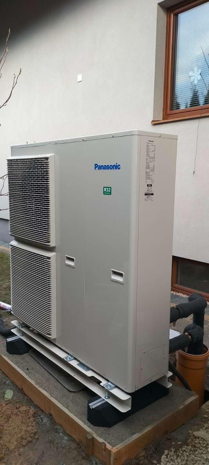 Zewnętrzna jednostka pompy ciepła Panasonic R32 zamontowana na betonowej podstawie z drewnianym obramowaniem, z widocznymi rurami w izolacji termicznej i oknem w tle.