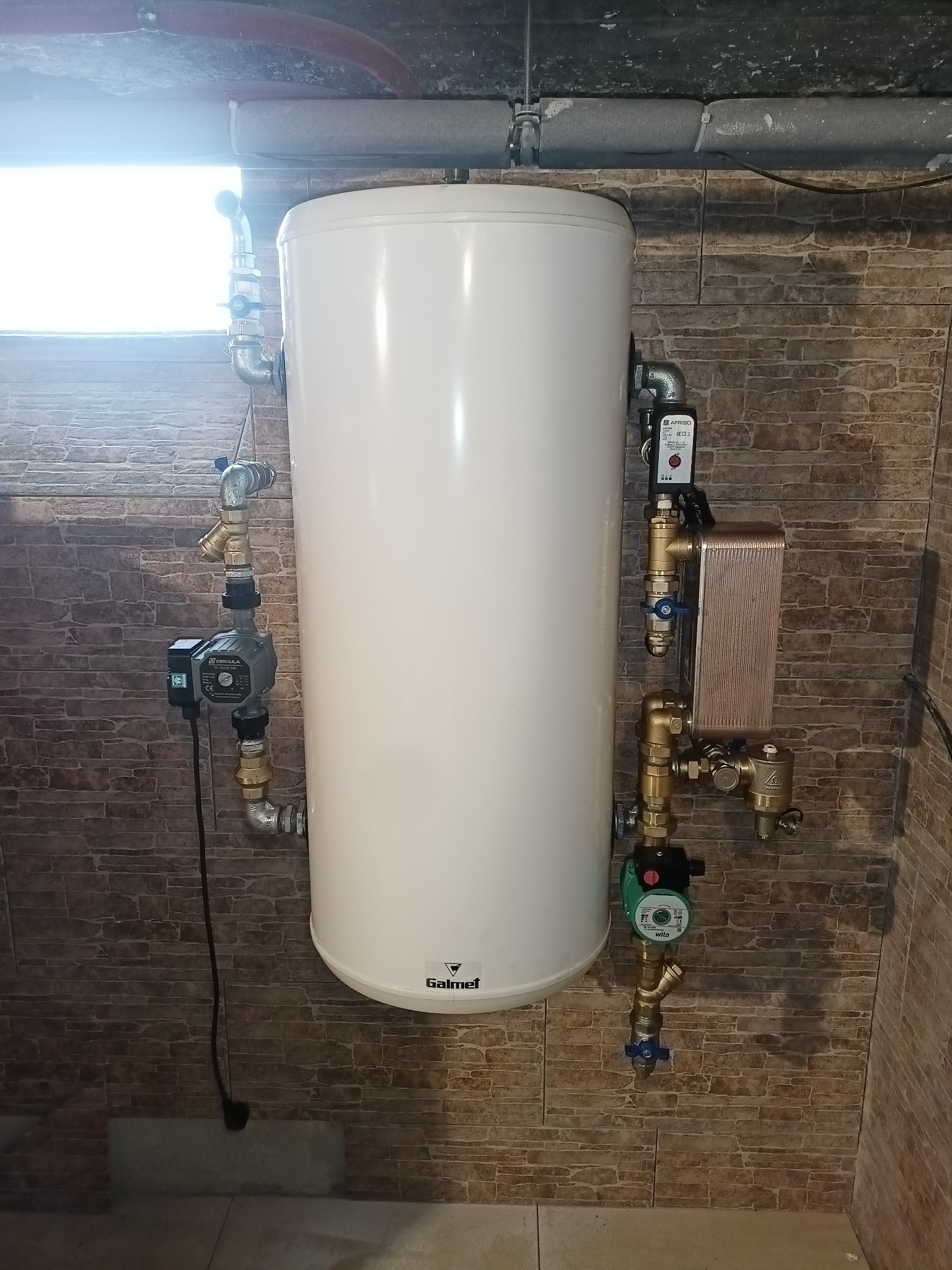 Biały, cylindryczny zbiornik na wodę Galmet z podłączonymi rurami, zaworami i pompami obiegowymi Wilo i Afriso, zamontowany na ścianie wyłożonej ceglastymi płytkami.