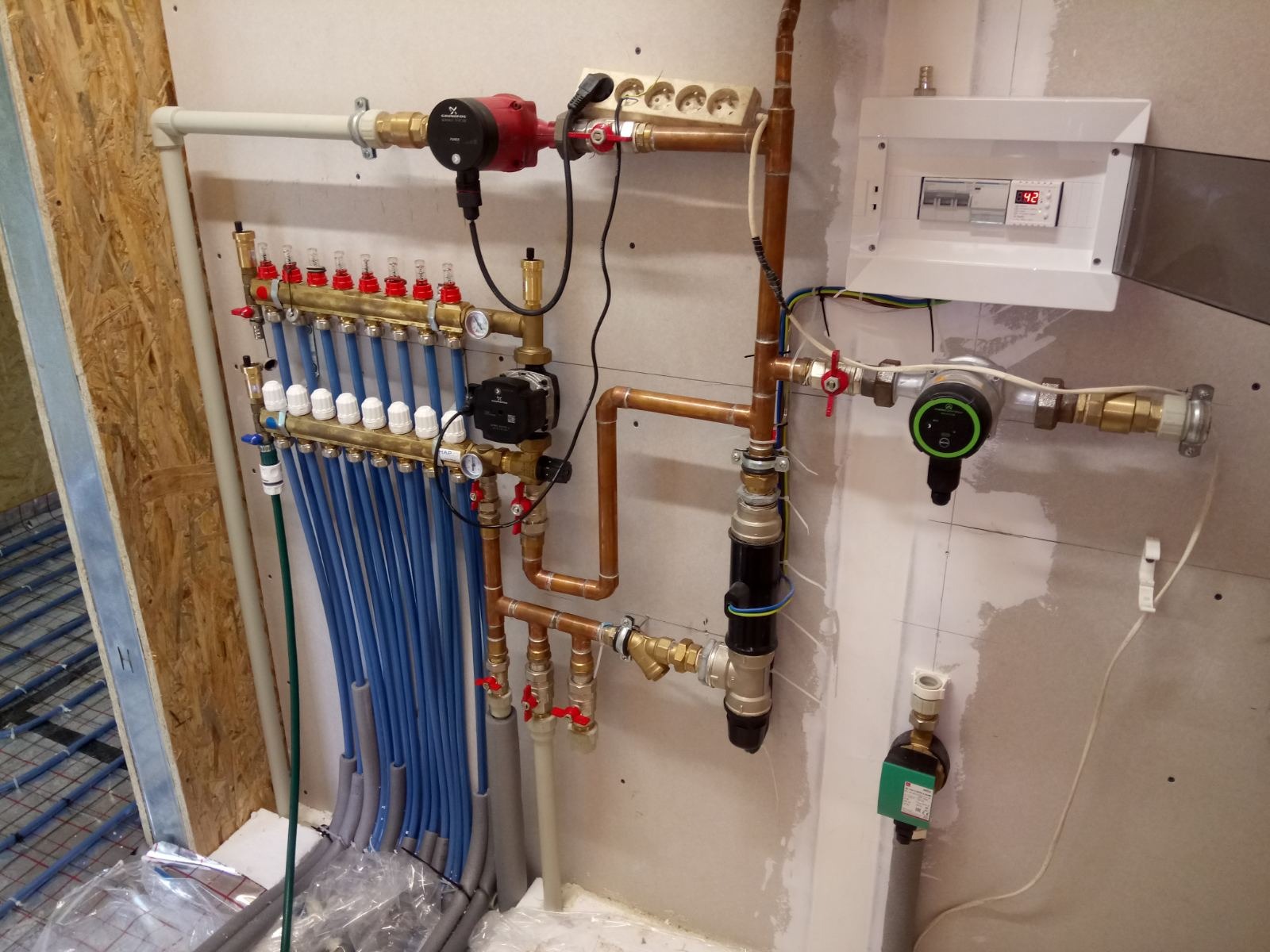 Rozdzielacz ogrzewania podłogowego z licznymi pętlami, pompami obiegowymi, zaworami i zabezpieczeniami, zamontowany na ścianie w surowym stanie, z widoczną instalacją elektryczną i skrzynką...