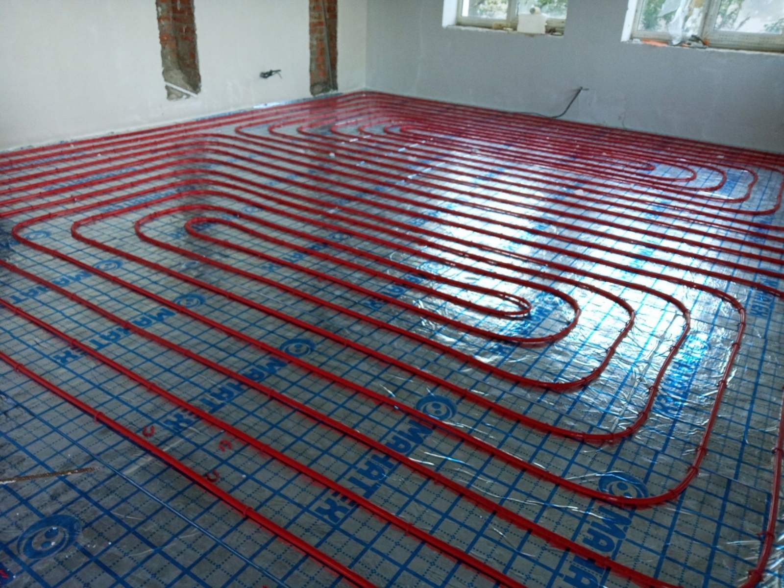 Czerwone rury ogrzewania podłogowego ułożone w pętle na folii z nadrukiem kratki w pomieszczeniu w stanie surowym, widoczne okna i fragmenty ścian bez tynku.