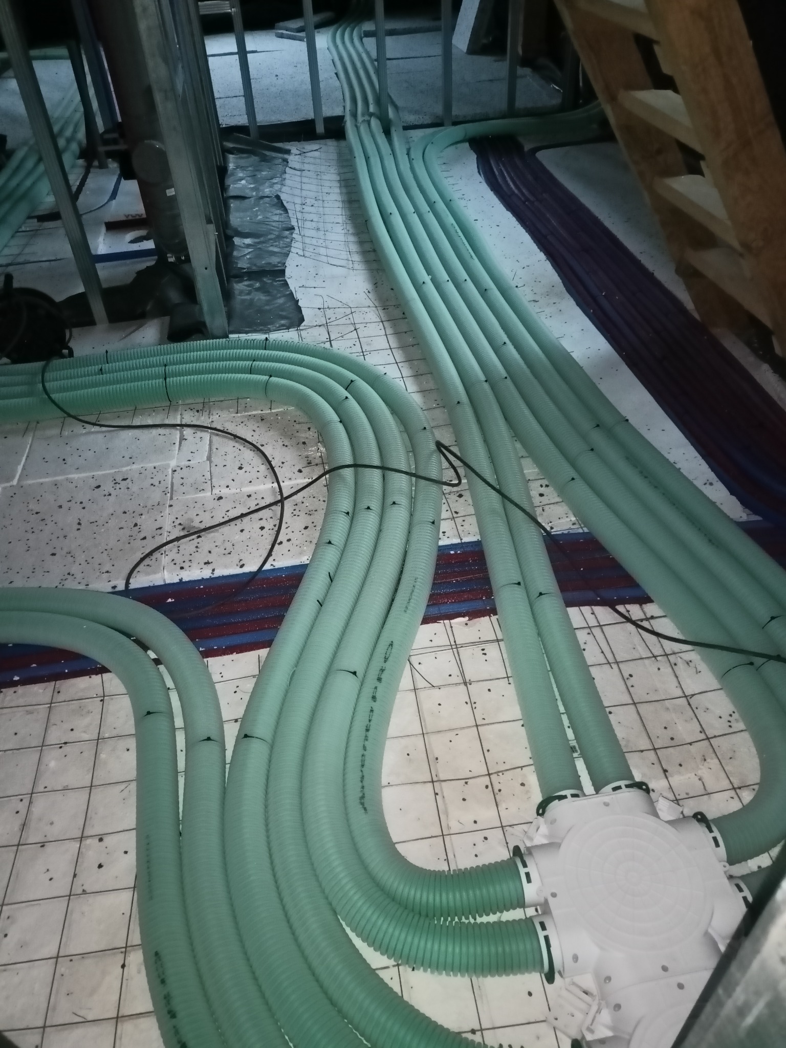 Rozprowadzenie instalacji ogrzewania podłogowego w peszlach koloru seledynowego, widoczna puszka rozdzielcza z licznymi wyjściami oraz fragmenty instalacji wodnej w kolorze czerwonym i niebieskim.