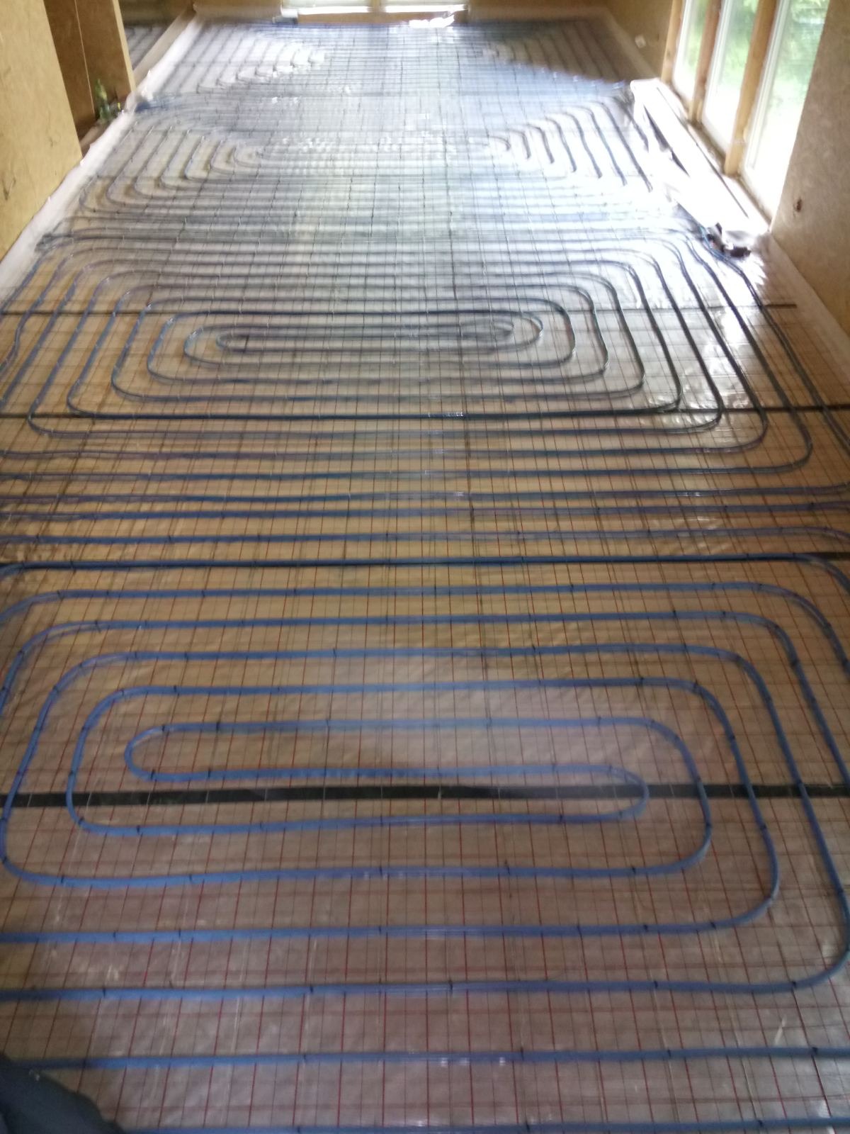 Równomiernie rozłożone, niebieskie rury ogrzewania podłogowego na metalowej siatce, widoczne okna w tle, surowy stan wykończenia wnętrza.