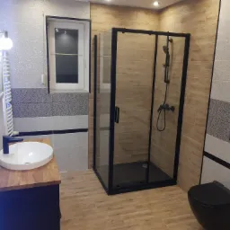 Wykończona łazienka z kabiną prysznicową w czarnej ramie, umywalką nablatową na drewnianym blacie i podwieszaną toaletą w kolorze czarnym, z oknem i grzejnikiem drabinkowym.