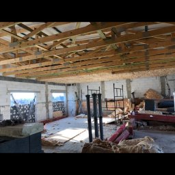 MODREPUS - Wnętrze w trakcie remontu: widoczne drewniane belki stropowe, ściany z białego pustaka, częściowo ocieplone ściany, rozłożona wełna mineralna, rusztowania oraz pionowe rury.