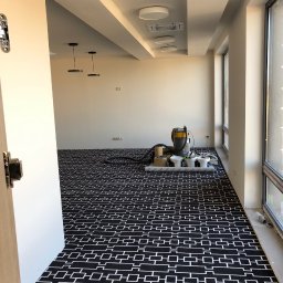 MODREPUS - Nowo położona czarno-biała wykładzina dywanowa we wzór geometryczny w jasnym pokoju, widoczna maszyna do czyszczenia dywanów i resztki materiałów wykończeniowych, ściany pomalowane na szaro.