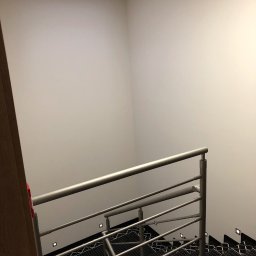 MODREPUS - Wnętrze klatki schodowej z metalową balustradą, czarno-białą wykładziną na schodach i kwadratowym oknem w suficie, oświetlającym białe ściany.