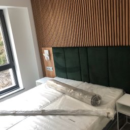 MODREPUS - Nowoczesna sypialnia z drewnianą ścianą z paneli, zielonym tapicerowanym zagłówkiem i łóżkiem z materacem w folii, obok okno i biała szafka nocna.