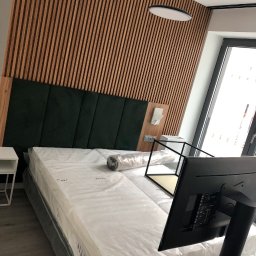 MODREPUS - Nowe łóżko z zielonym wezgłowiem i drewnianą ścianą dekoracyjną w pokoju, materac jeszcze w folii ochronnej.