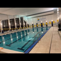 MODREPUS - Wnętrze krytego basenu z niebieską wodą, drabinkami ze stali nierdzewnej, leżakami i dekoracyjnymi chorągiewkami w kolorach żółtym i niebieskim, widok na logo 'interferie.pl' na ścianie.