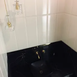 Czarna umywalka nablatowa o kwadratowym kształcie z armaturą w kolorze złotym na blacie z czarnego marmuru z żyłami, białe płytki na ścianie i wiszące lampy dekoracyjne w kształcie flakon&oacute;w.