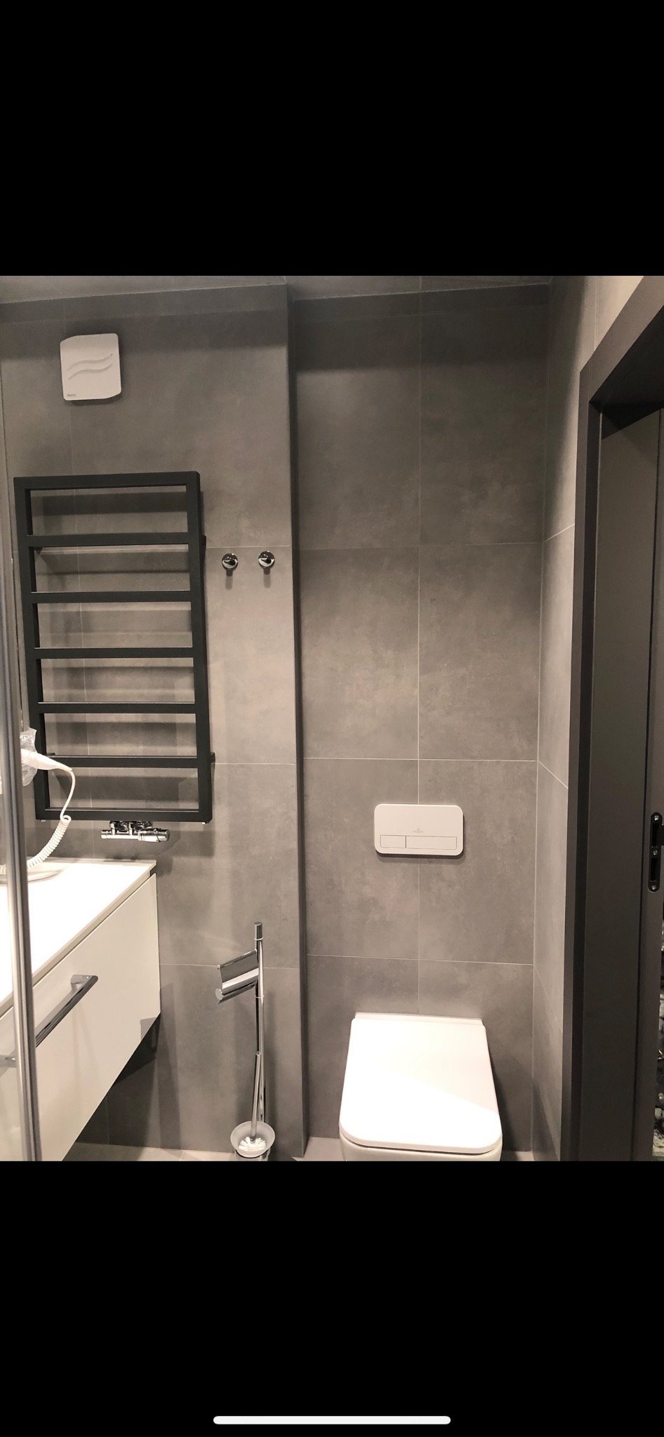 Nowoczesna łazienka z szarymi płytkami, wiszącą toaletą, grzejnikiem drabinkowym i białą szafką umywalkową.