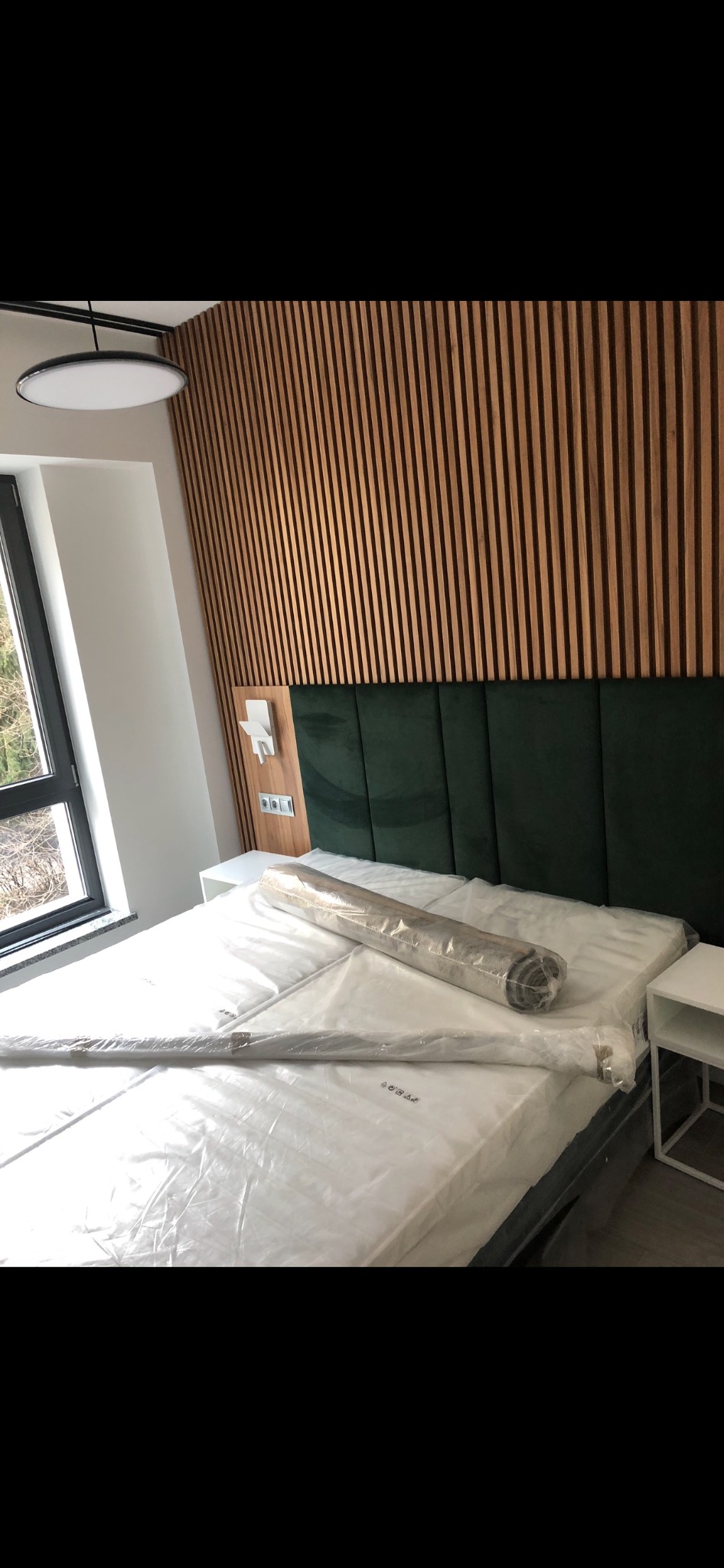 Nowoczesna sypialnia z drewnianą ścianą z paneli, zielonym tapicerowanym zagłówkiem i łóżkiem z materacem w folii, obok okno i biała szafka nocna.