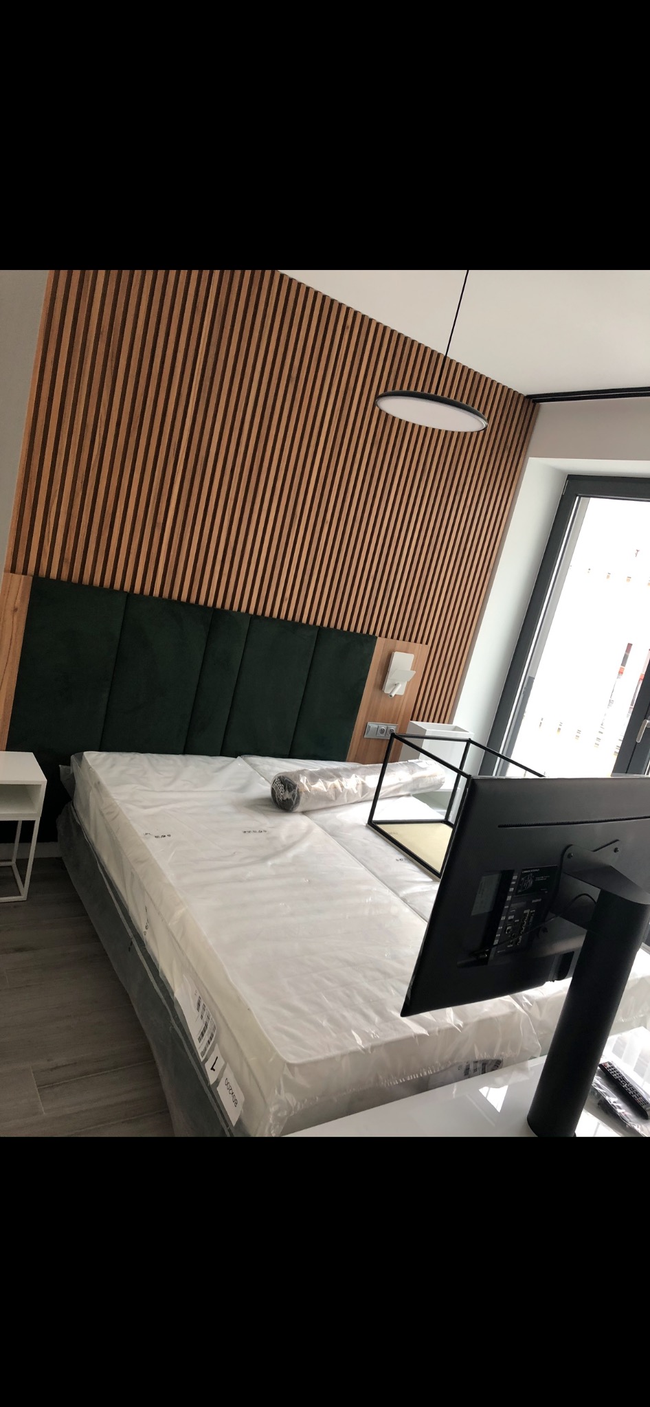 Nowe łóżko z zielonym wezgłowiem i drewnianą ścianą dekoracyjną w pokoju, materac jeszcze w folii ochronnej.