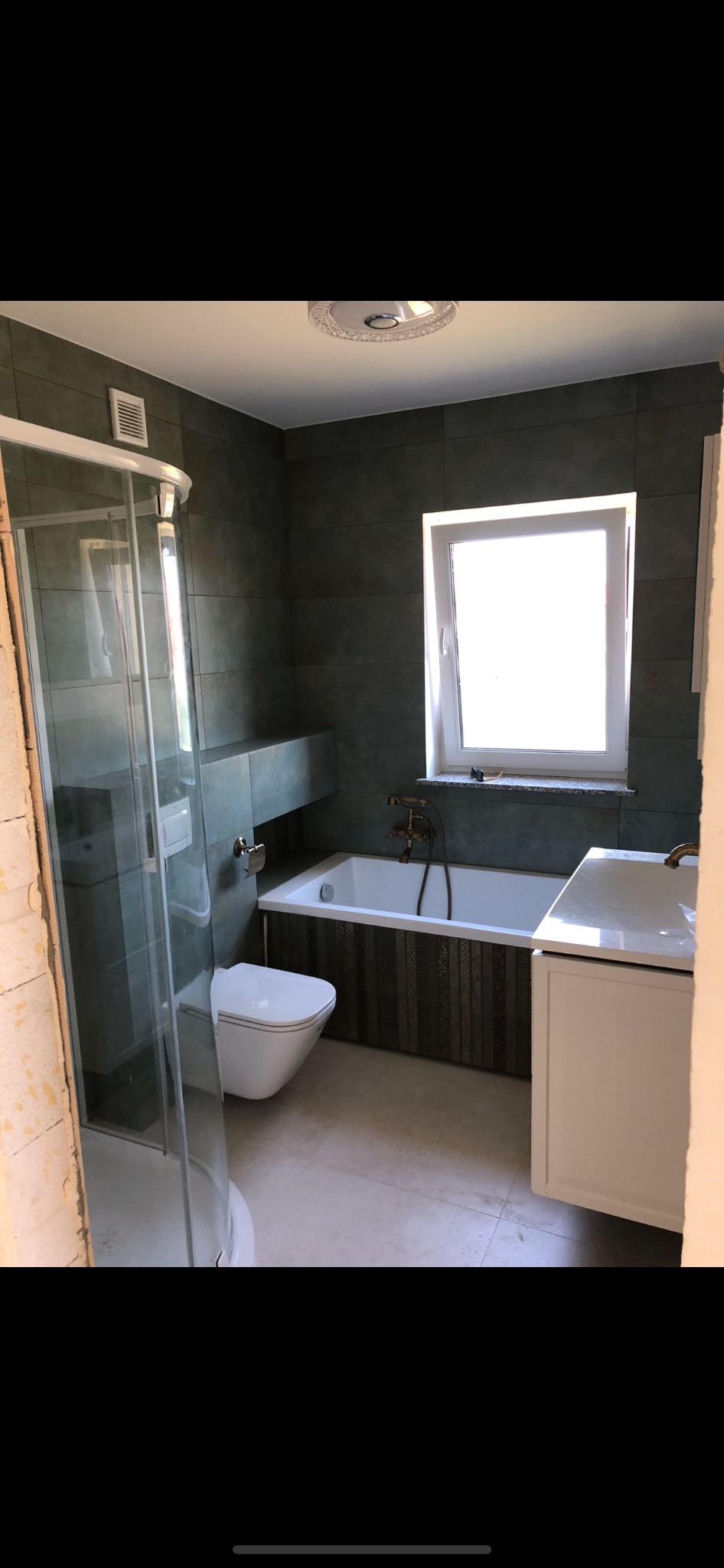 Nowoczesna łazienka z kabiną prysznicową, toaletą, wanną obudowaną dekoracyjnymi panelami i umywalką z szafką, wykończona w ciemnych płytkach z jasną podłogą.