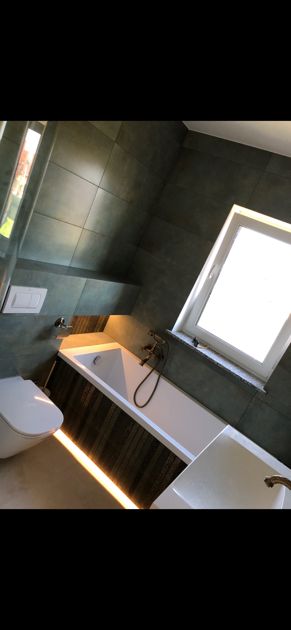 Wykończona łazienka z ciemnymi płytkami, podświetlaną wanną, białą armaturą i oknem z marmurowym parapetem.