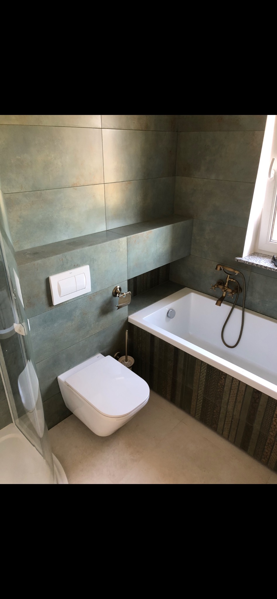 Nowoczesna łazienka z wiszącą toaletą, wanną z ozdobną obudową i baterią w stylu retro, wykończona płytkami w odcieniach zieleni i beżu.