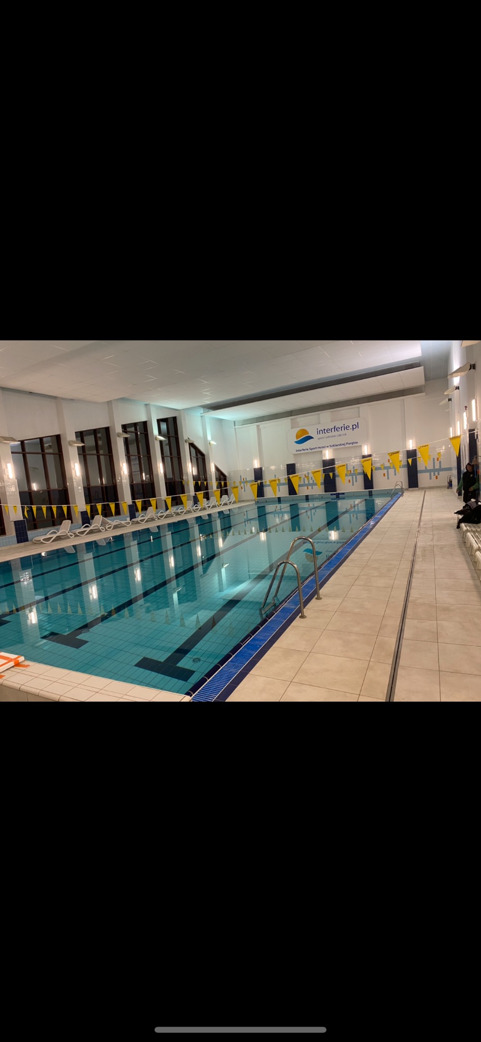 Wnętrze krytego basenu z niebieską wodą, drabinkami ze stali nierdzewnej, leżakami i dekoracyjnymi chorągiewkami w kolorach żółtym i niebieskim, widok na logo 'interferie.pl' na ścianie.