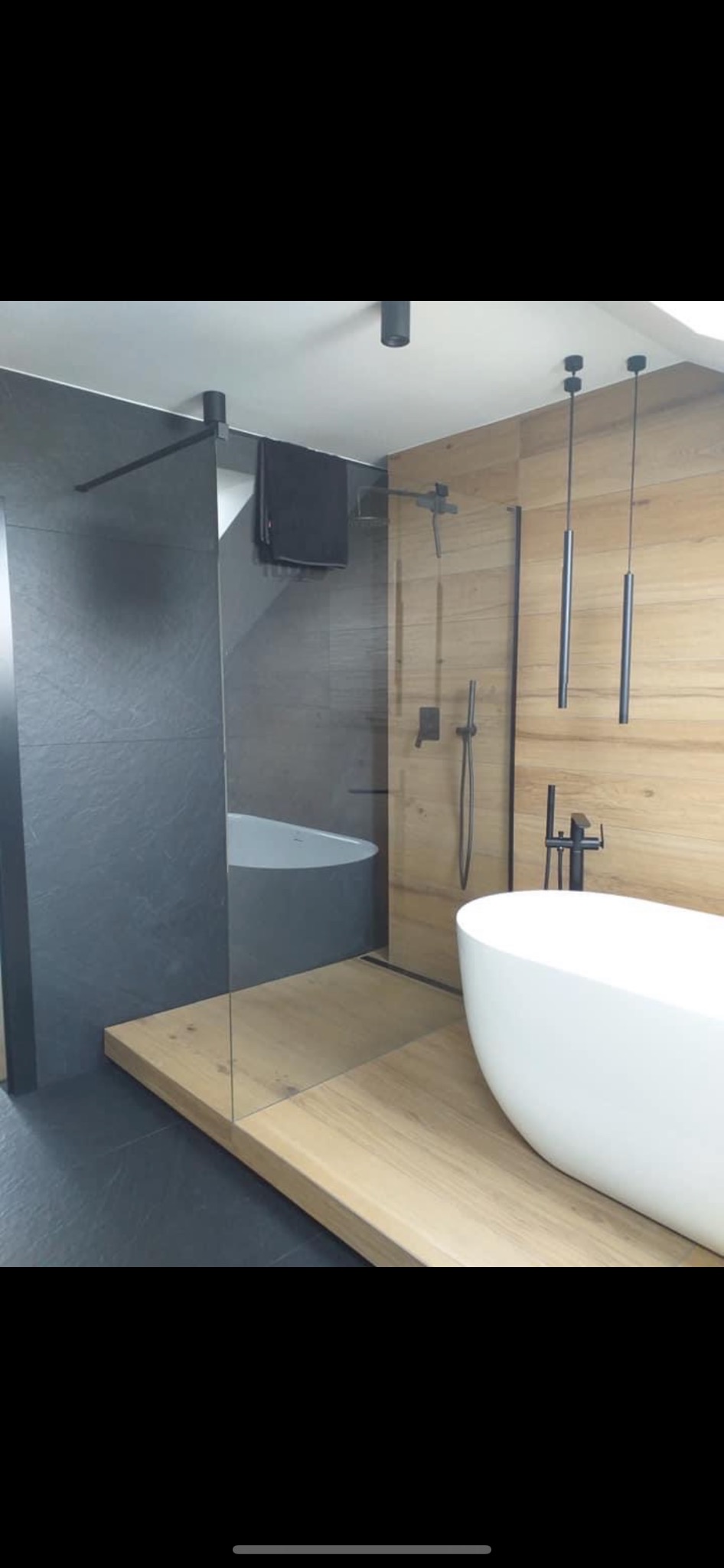 Nowoczesna łazienka z drewnianą podłogą, wolnostojącą białą wanną, szklaną kabiną prysznicową i czarnymi elementami armatury oraz oświetlenia.