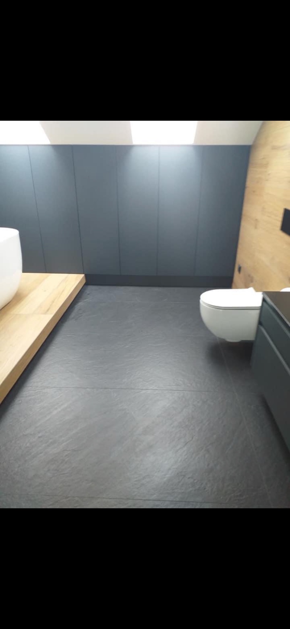 Nowoczesna łazienka z szarą płytką podłogową, drewnianym blatem pod umywalką, białą toaletą i szarą zabudową ścienną.