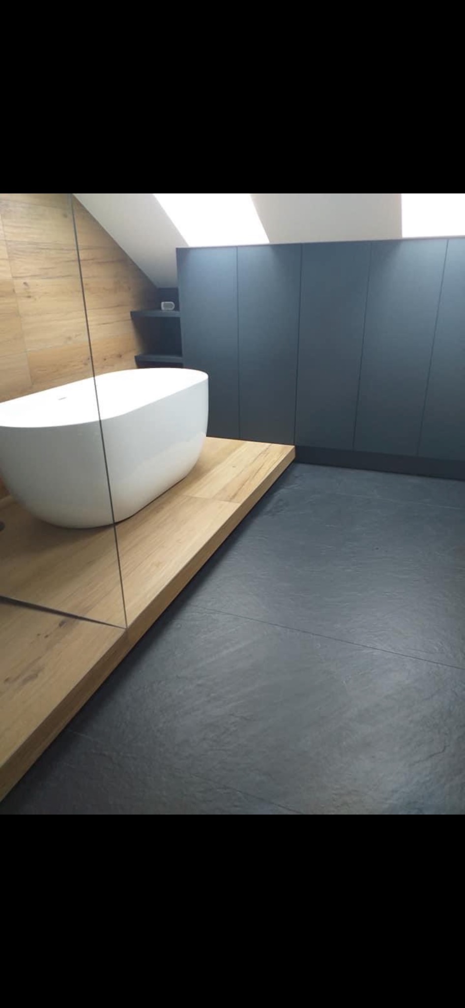 Nowoczesna łazienka z wolnostojącą białą wanną na drewnianym podeście, szara podłoga i zabudowa meblowa w kolorze antracytowym pod skośnym sufitem.