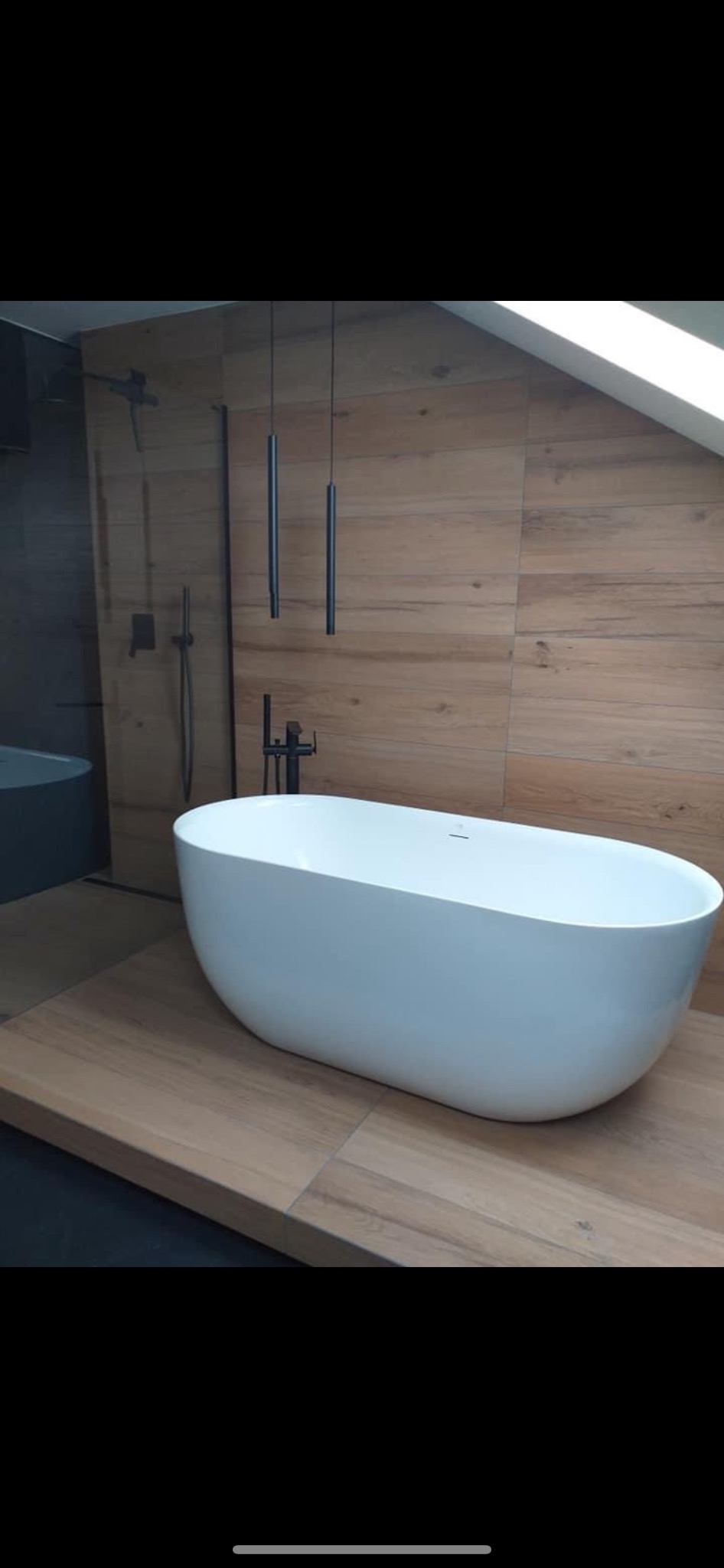 Wolnostojąca, owalna wanna w nowoczesnej łazience na poddaszu, z drewnopodobnymi płytkami na ścianach i podłodze, czarna armatura i minimalistyczne lampy wiszące.