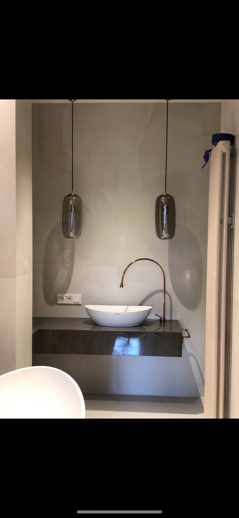 Nowoczesna łazienka z owalną umywalką nablatową, miedzianą baterią i dwiema szklanymi lampami wiszącymi, widoczny fragment wanny.