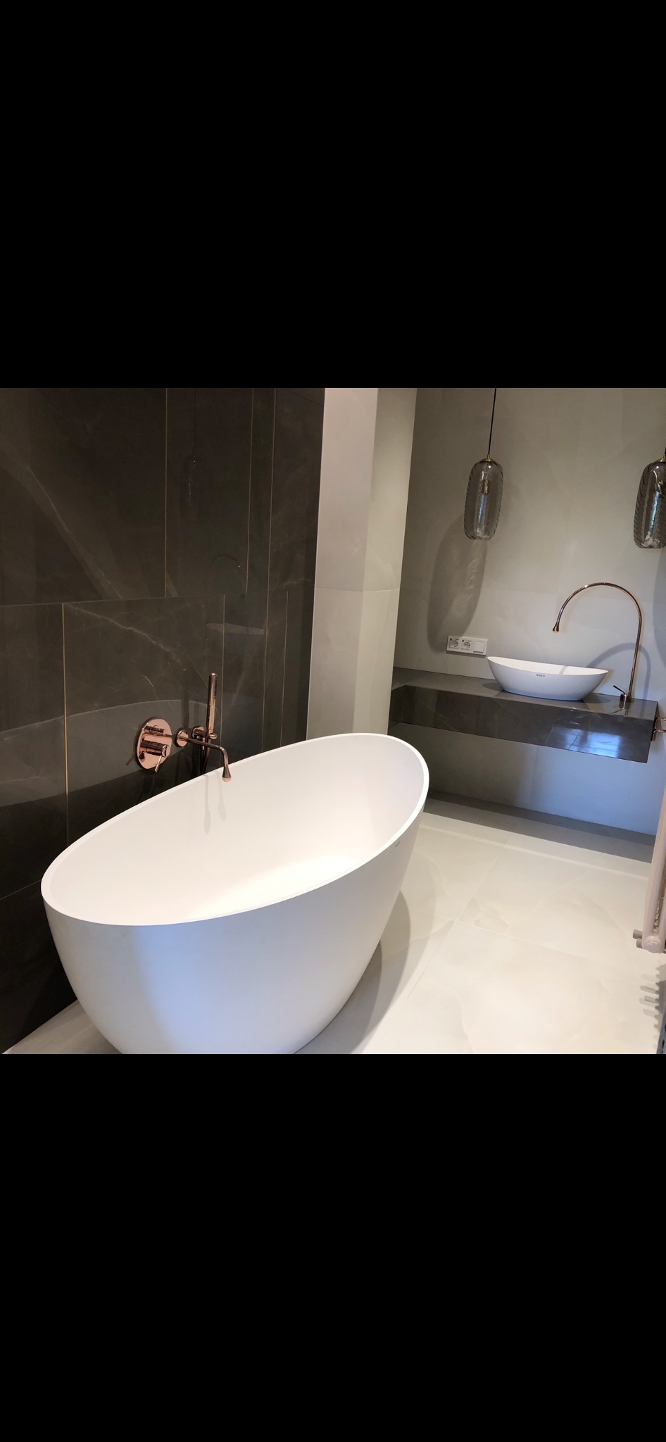 Nowoczesna łazienka z wolnostojącą białą wanną o owalnym kształcie, armaturą w kolorze różowego złota i umywalką nablatową na marmurowym blacie, oświetlona wiszącymi lampami.