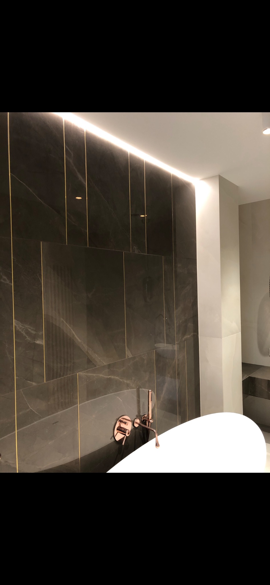 Luksusowa łazienka z ciemnymi płytkami marmuropodobnymi, złotymi wstawkami, podświetleniem LED i armaturą w kolorze różowego złota nad białą wanną wolnostojącą.