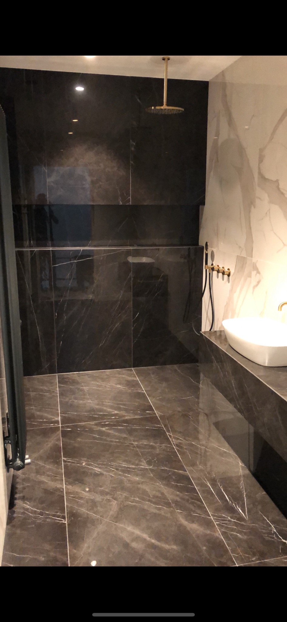 Nowoczesna łazienka z ciemnymi marmurowymi płytkami na ścianach i podłodze, złota deszczownica i bateria umywalkowa, biała umywalka nablatowa.