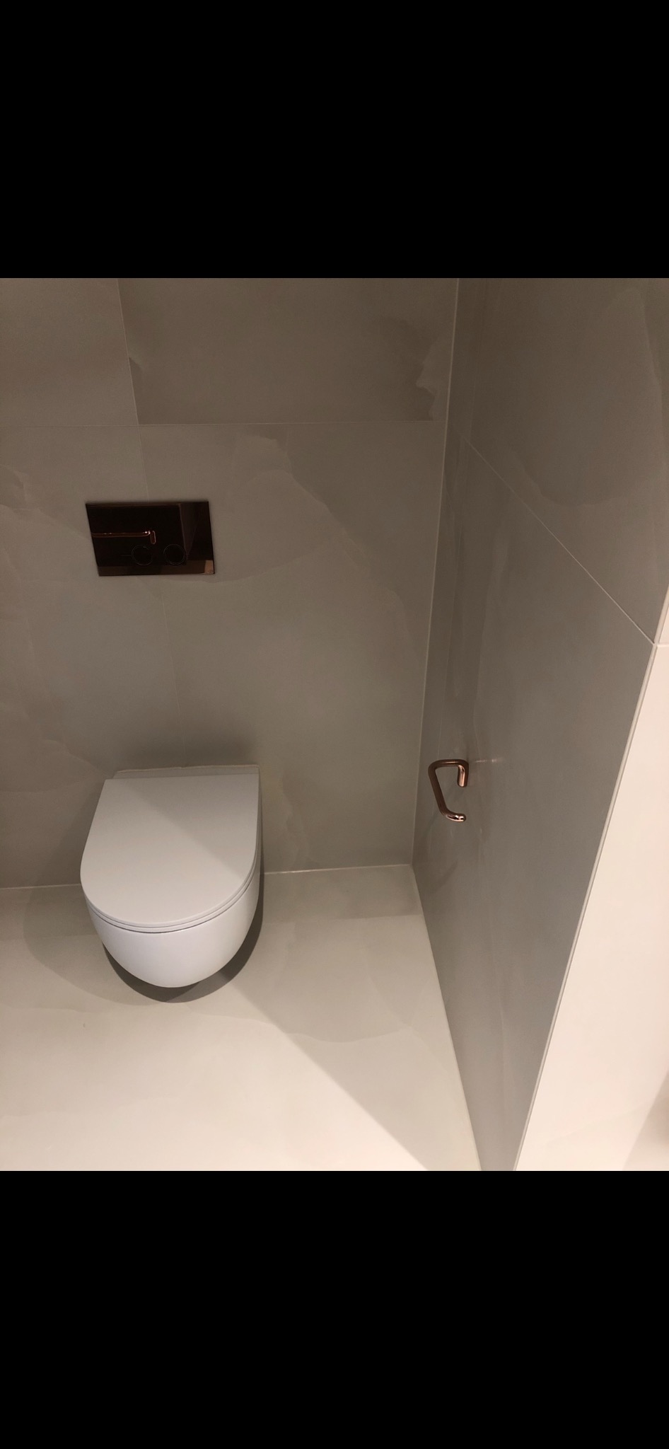 Nowoczesna toaleta podwieszana z chromowanym przyciskiem spłukującym i uchwytem na papier toaletowy w łazience wykończonej szarymi płytkami imitującymi marmur.