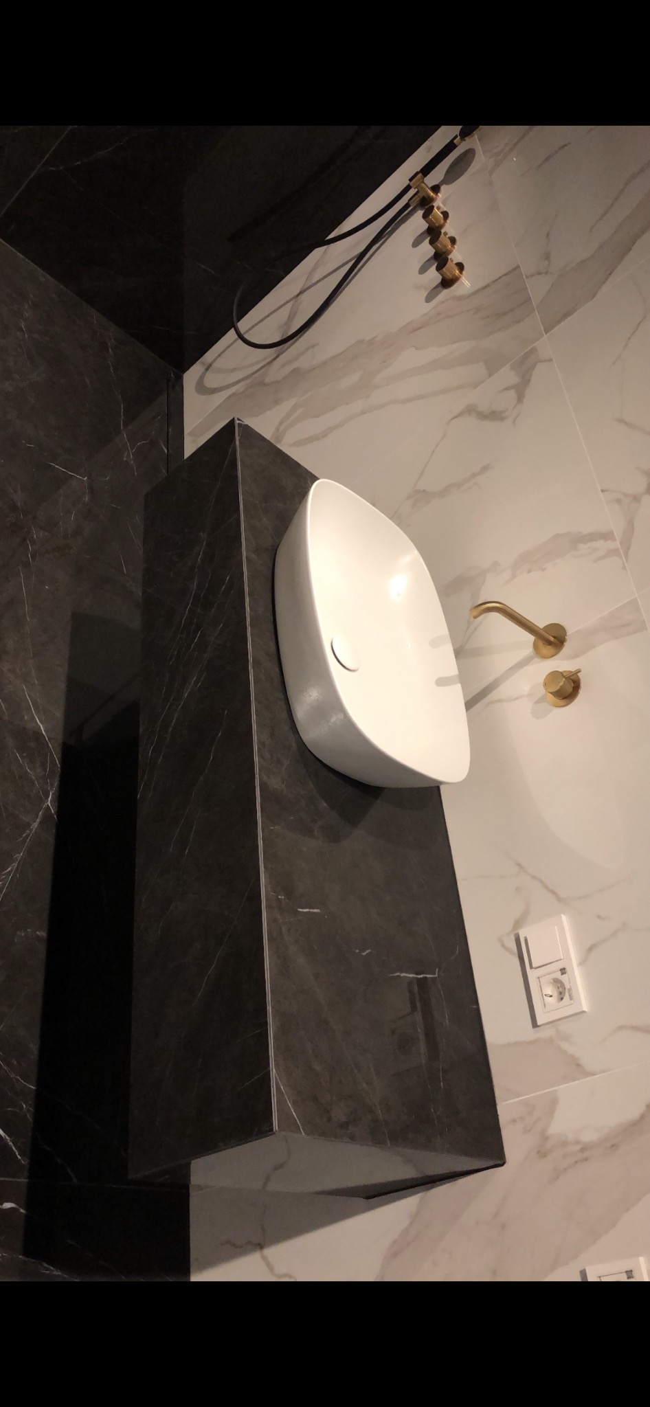 Umywalka nablatowa o owalnym kształcie, zamontowana na postumencie z ciemnego marmuru, z baterią w kolorze szczotkowanego złota i marmurową ścianą w tle.