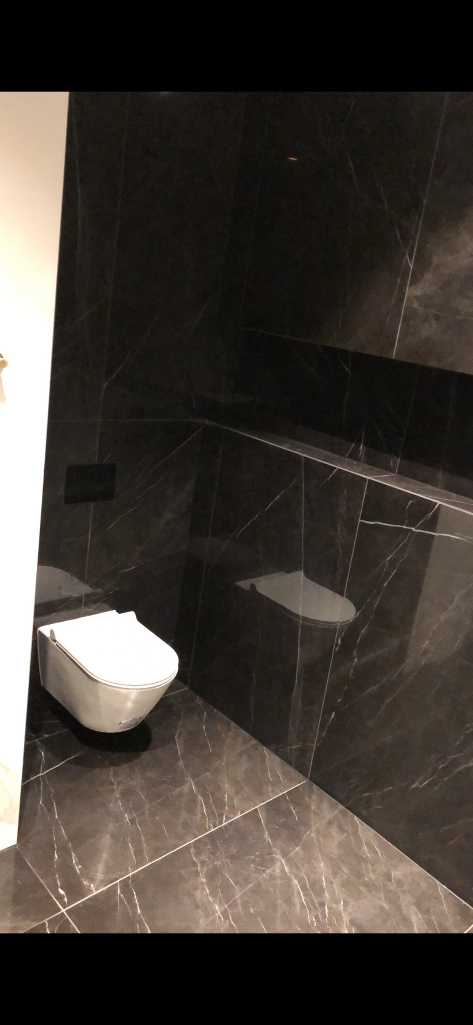 Nowoczesna łazienka z wiszącą toaletą i ciemnymi płytkami imitującymi marmur, z wnęką w ścianie wykończoną tym samym materiałem.