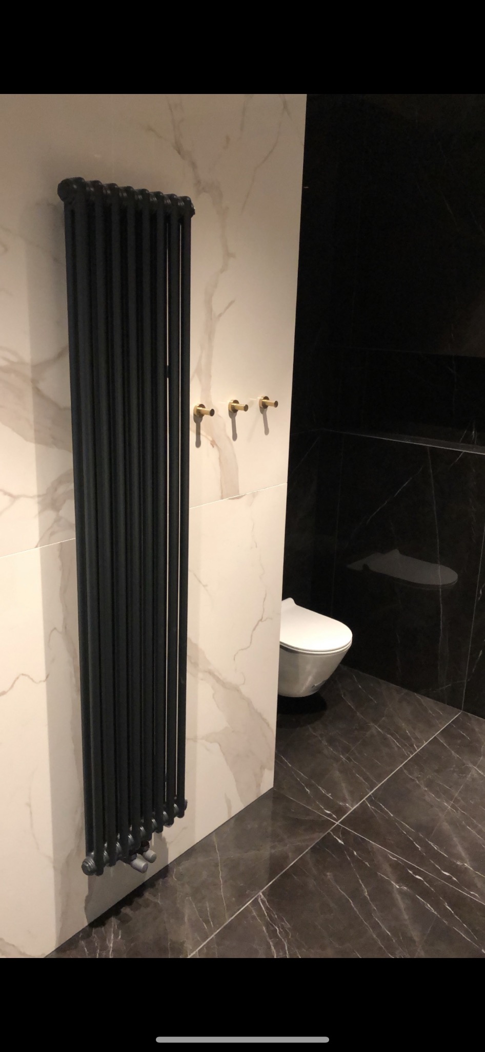 Czarny, żeberkowy grzejnik łazienkowy na białej ścianie imitującej marmur, obok trzy złote wieszaki i toaleta na czarnej ścianie z marmurowym wzorem, podłoga z ciemnego marmuru.