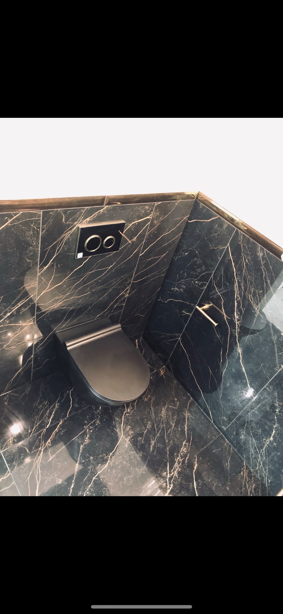 Nowoczesna toaleta podwieszana w kolorze grafitowym, zamontowana w rogu łazienki wykończonej czarnymi płytkami imitującymi marmur ze złotymi żyłami; widoczny panel spłukujący i fragment złotego...