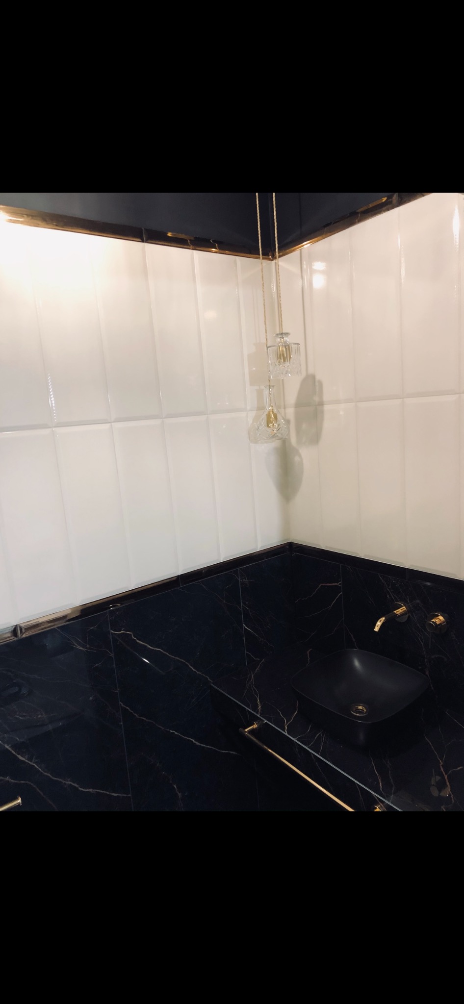 Nowoczesna łazienka w Sycowie z czarnym blatem z marmurowym wzorem, białe kafelki na ścianach, czarna umywalka i złote detale, w tym bateria i wieszak na ręczniki oraz lampa wisząca.