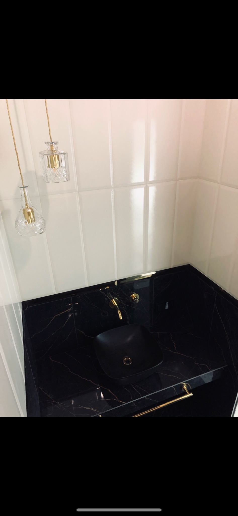 Czarna umywalka nablatowa o kwadratowym kształcie z armaturą w kolorze złotym na blacie z czarnego marmuru z żyłami, białe płytki na ścianie i wiszące lampy dekoracyjne w kształcie flakonów.
