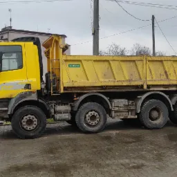 Żółta ciężarówka wywrotka firmy SKIBICKI z widocznymi śladami użytkowania na mokrym placu, z boku widoczna koparka, w tle budynki mieszkalne i słupy energetyczne.