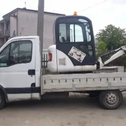 Biały samochód dostawczy z platformą transportową przewożący małą, białą koparkę z naklejką firmy Bud-Kop Staniek, na tle zabudowy jednorodzinnej.