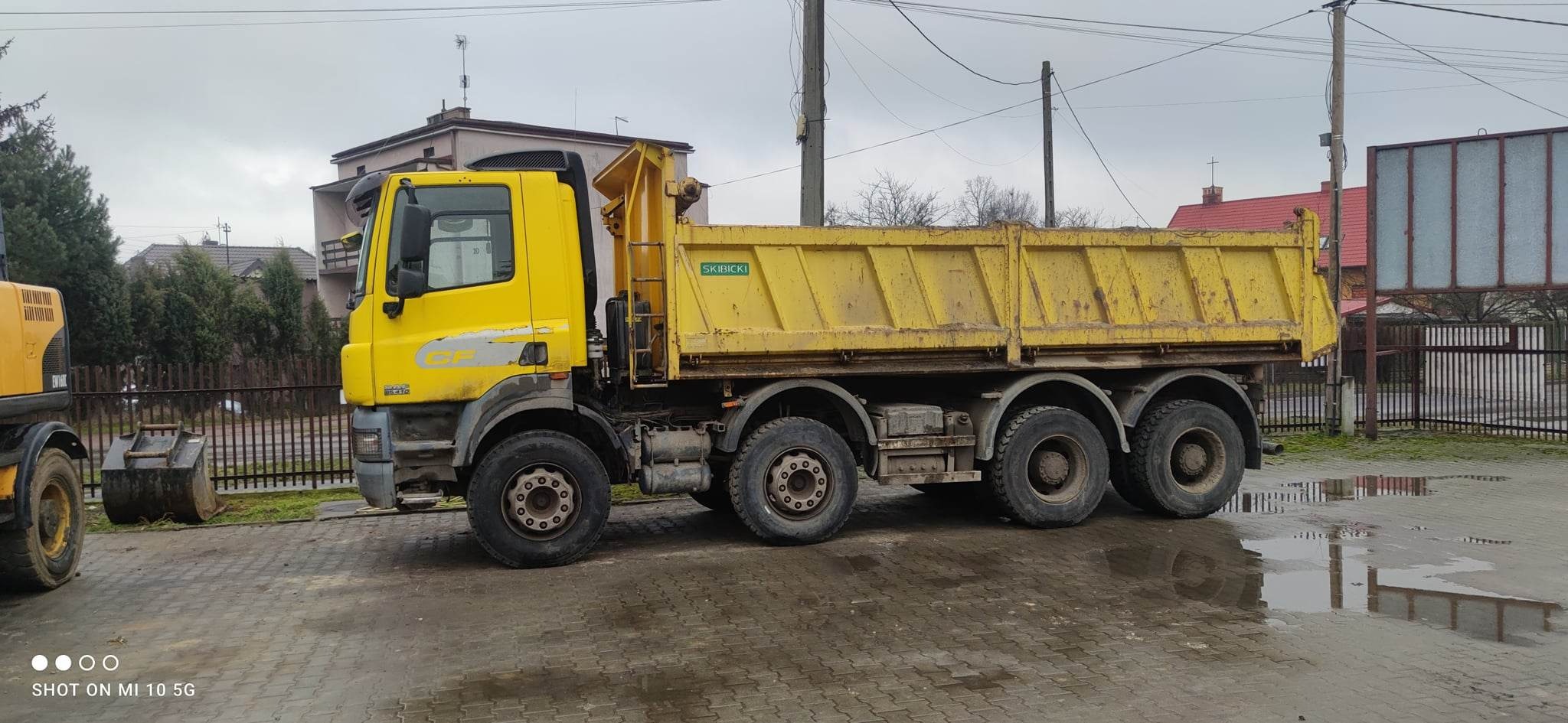 Żółta ciężarówka wywrotka firmy SKIBICKI z widocznymi śladami użytkowania na mokrym placu, z boku widoczna koparka, w tle budynki mieszkalne i słupy energetyczne.