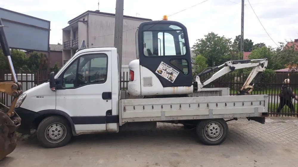 Biały samochód dostawczy z platformą transportową przewożący małą, białą koparkę z naklejką firmy Bud-Kop Staniek, na tle zabudowy jednorodzinnej.