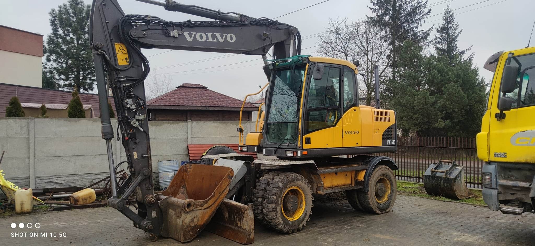 Żółta koparka kołowa Volvo EW160C z brudną łyżką, zaparkowana na placu budowy z widocznym w tle betonowym ogrodzeniem i budynkiem z czerwonym dachem.